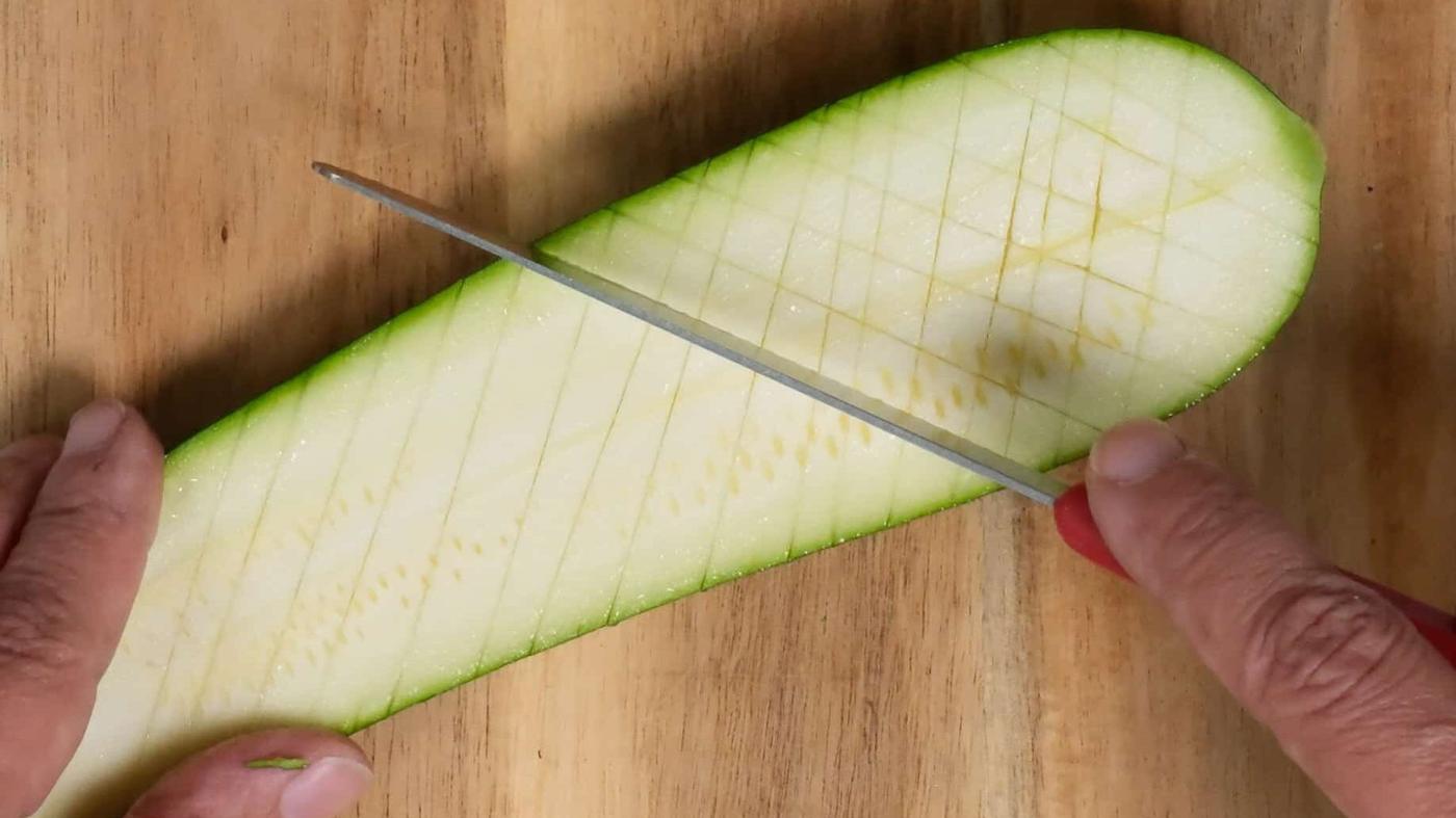 scoring a halved zucchini with a knife in a crosshatch pattern