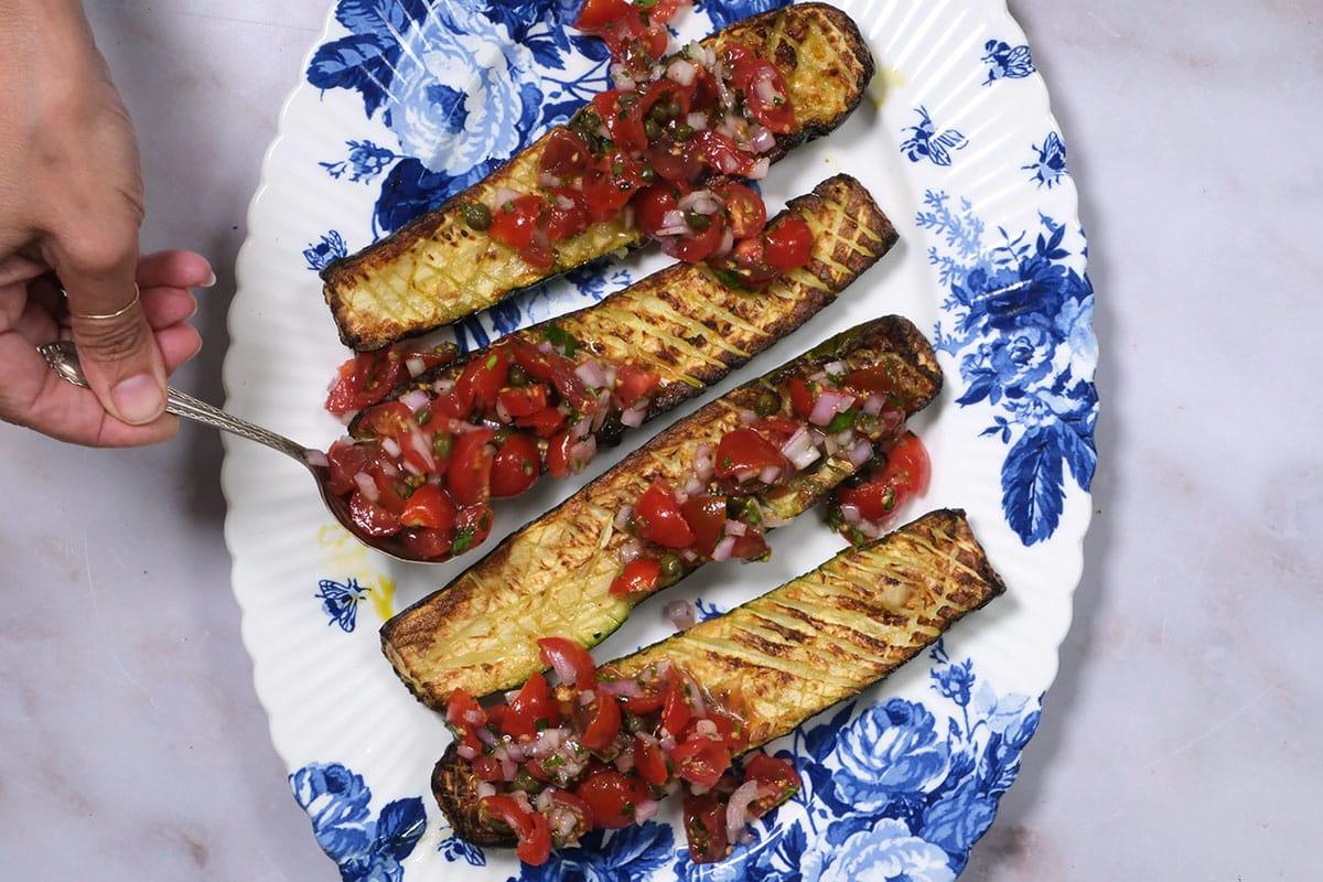 spooning tomato sauce over roasted zucchini on a floral platter