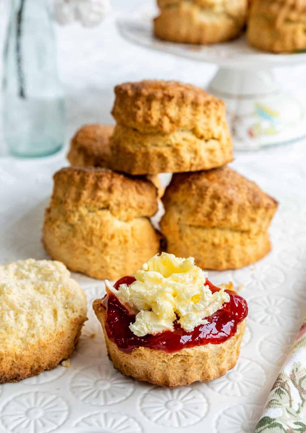 A close-up of a scone split in half, topped with red jam and clotted cream, in front of a stack of plain Air Fryer Scones on a white patterned surface. A glass bottle and plate of more scones in air fryer style are in the background.