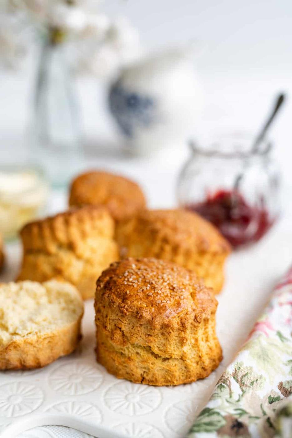 Golden brown air fryer scones are arranged on a white surface, with one scone split open. In the background, there’s a jar of jam, a floral napkin, and blurred decorative items