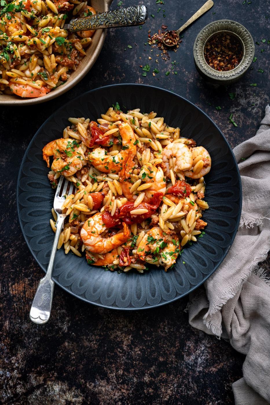 A black plate filled with Shrimp orzo recipe—featuring orzo pasta, shrimp, sun-dried tomatoes, and herbs—sits on a dark surface with a fork, beige napkin nearby, and a bowl of chili flakes and another serving dish in the background.
