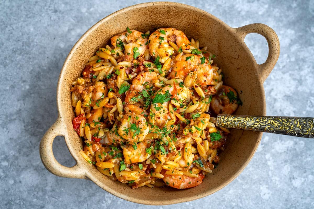 A tan ceramic bowl filled with cooked shrimp, orzo pasta, tomato pieces, and fresh chopped herbs showcases a delicious shrimp orzo recipe, with a decorative metal spoon resting inside the dish on a gray textured surface.