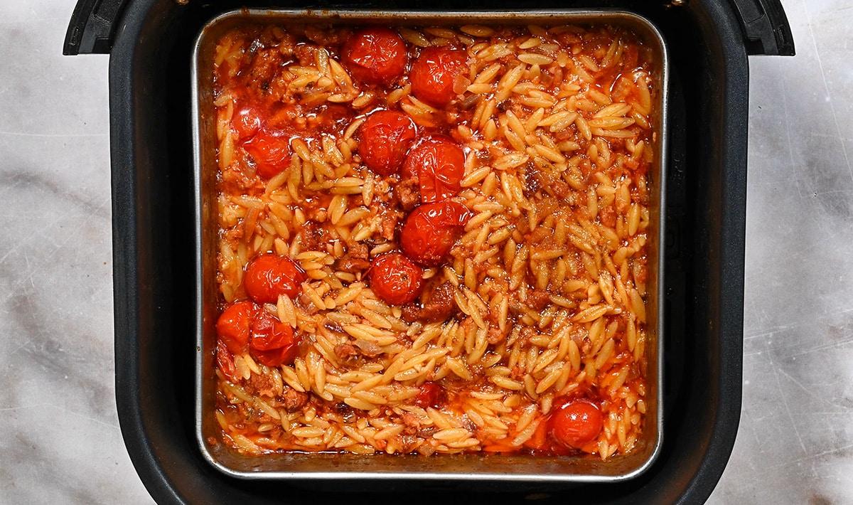 A square baking dish filled with cooked orzo pasta, cherry tomatoes, and a tomato-based sauce sits inside an air fryer on a marble countertop—a perfect Shrimp orzo recipe waiting to be enjoyed.
