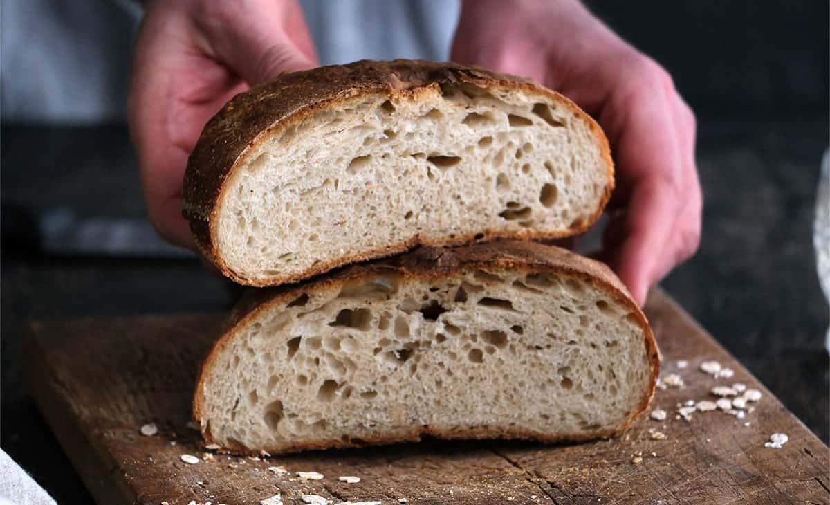 A person holds a sliced round loaf of rustic air fryer sourdough bread, showing its airy crumb and golden-brown crust, on a wooden cutting board.