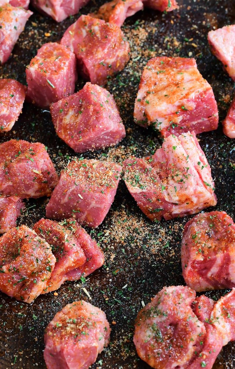 Chunks of raw beef coated with seasoning spices and herbs are spread out on a dark surface, ready to be cooked into delicious Air Fryer Steak Bites.