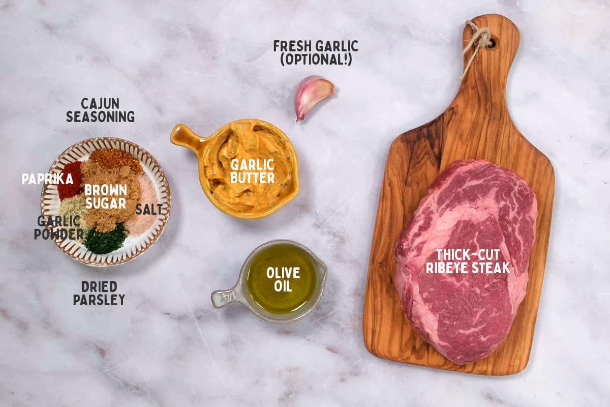 A thick-cut ribeye steak on a wooden board. Nearby are a clove of garlic, a bowl of garlic butter, and spices like paprika, Cajun seasoning, brown sugar, etc. Perfect for transforming into juicy steak bites in an air fryer. Fresh garlic is optional.