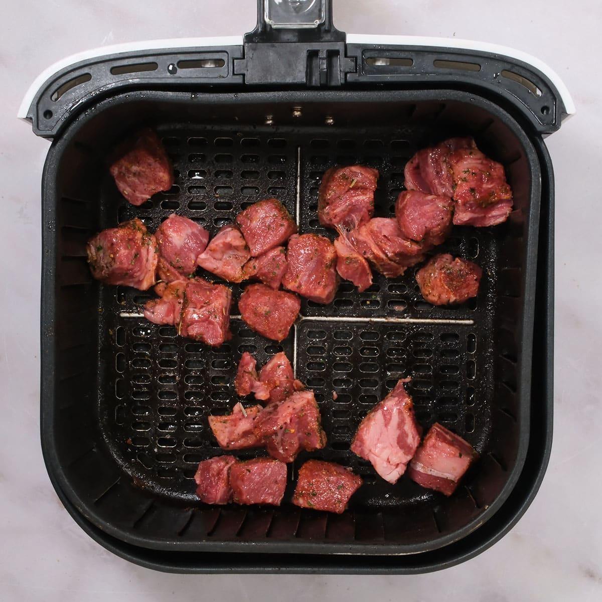 Seasoned cubes of raw steak, perfect for Air Fryer Steak Bites, are spread out inside the basket of an air fryer. The black basket features a grated bottom for even cooking.