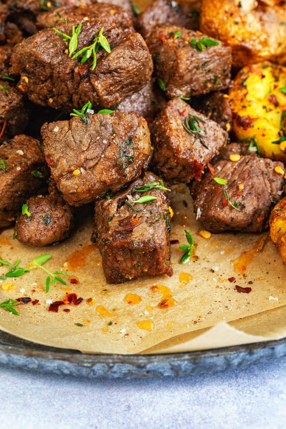 Close-up of juicy Air Fryer Steak Bites garnished with fresh herbs and red pepper flakes, served on parchment paper with a piece of roasted potato in the background.