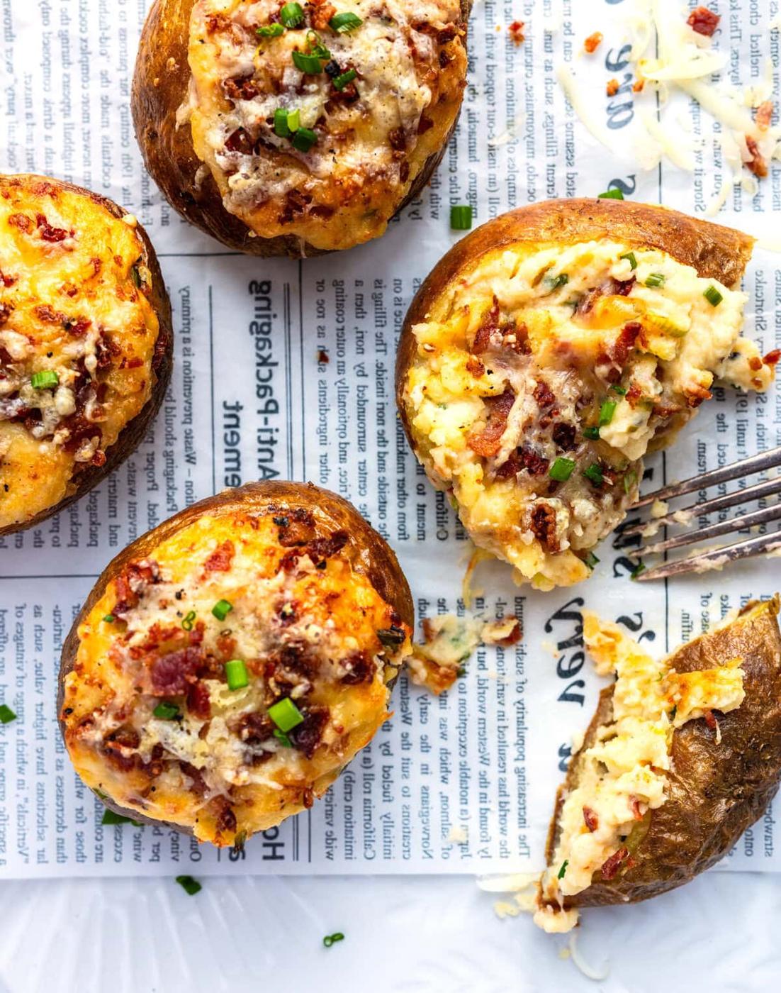 Four twice-baked potatoes with cheese, bacon bits, and chopped chives are served on newspaper. One potato is cut open with a fork, showing a creamy, cheesy filling. Grated cheese is scattered around.