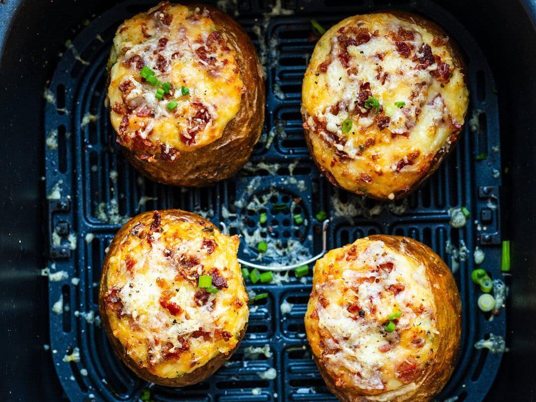 Four twice-baked potatoes topped with melted cheese, bacon bits, and chopped green onions are arranged inside an air fryer basket.