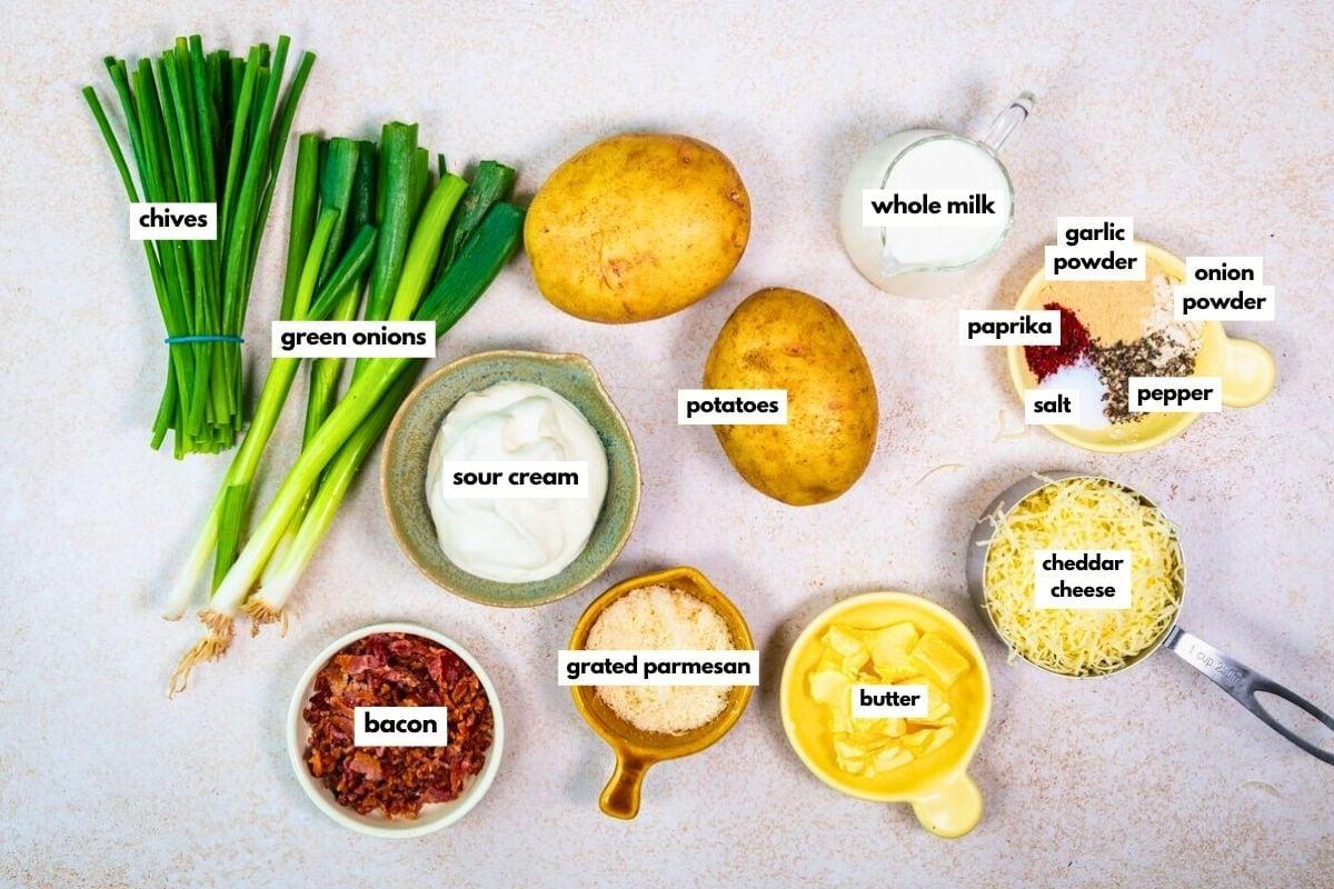 Labeled ingredients for mashed potatoes are arranged on a light surface: chives, green onions, sour cream, potatoes, whole milk, grated parmesan, bacon, butter, cheddar cheese, paprika, garlic powder, onion powder, salt, and pepper.