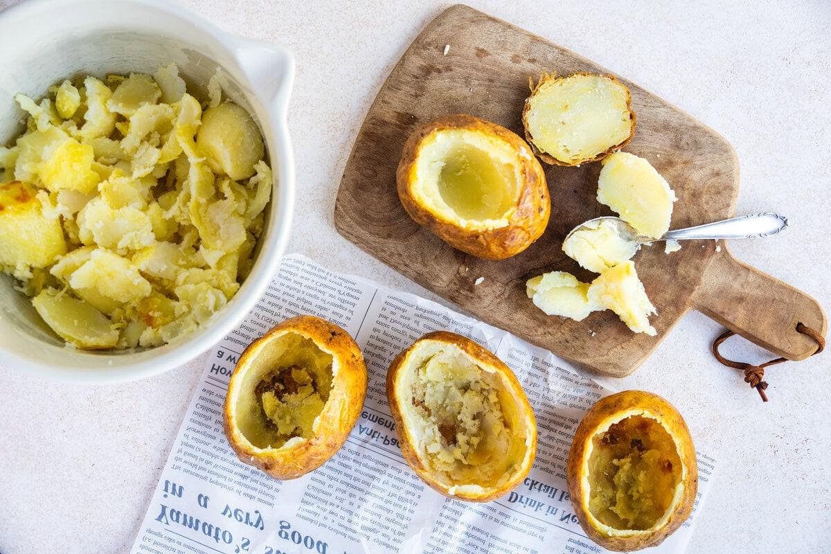 Three hollowed baked potatoes on newspaper, baked potato tops and scooped potato flesh on a wooden cutting board, and a bowl filled with mashed potato, with a spoon nearby.