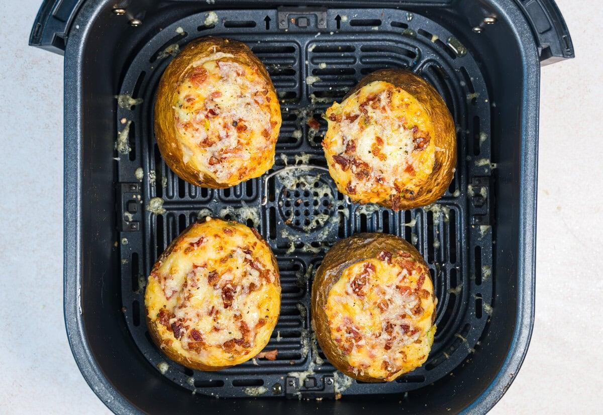 Four stuffed baked potatoes with melted cheese and bacon sit in the basket of an air fryer, viewed from above. The potatoes have golden, crispy tops and are arranged in a square formation.