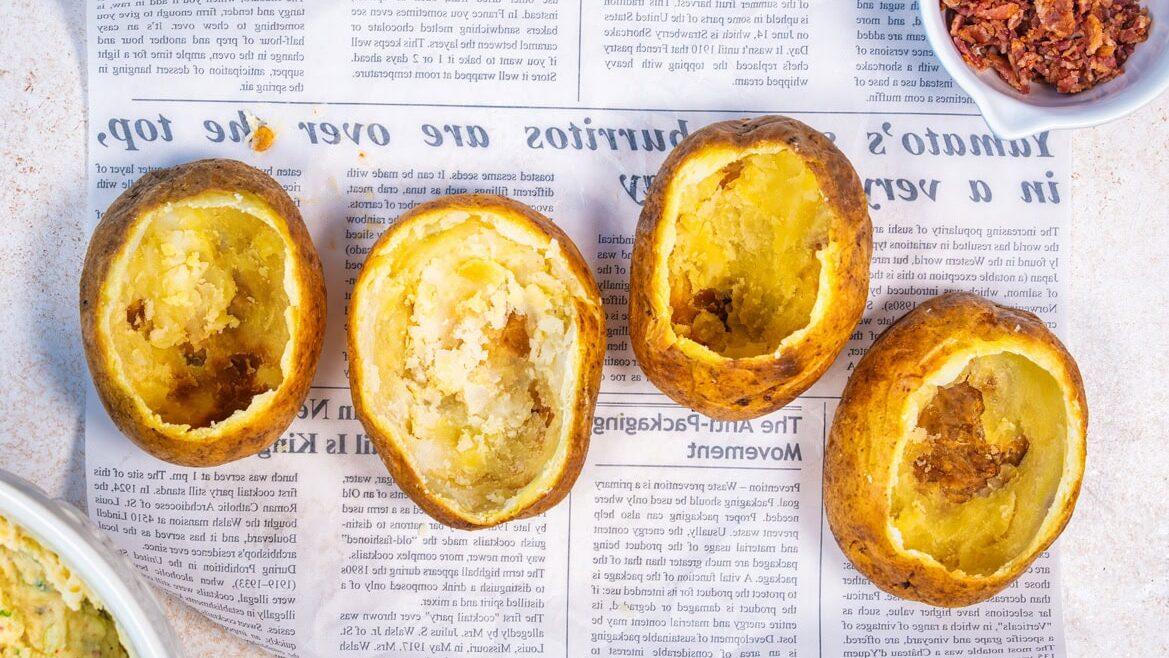 Four baked potato skins with their insides scooped out are lined up on newspaper, with a bowl of crumbled bacon bits and a dish of mashed potato filling nearby.