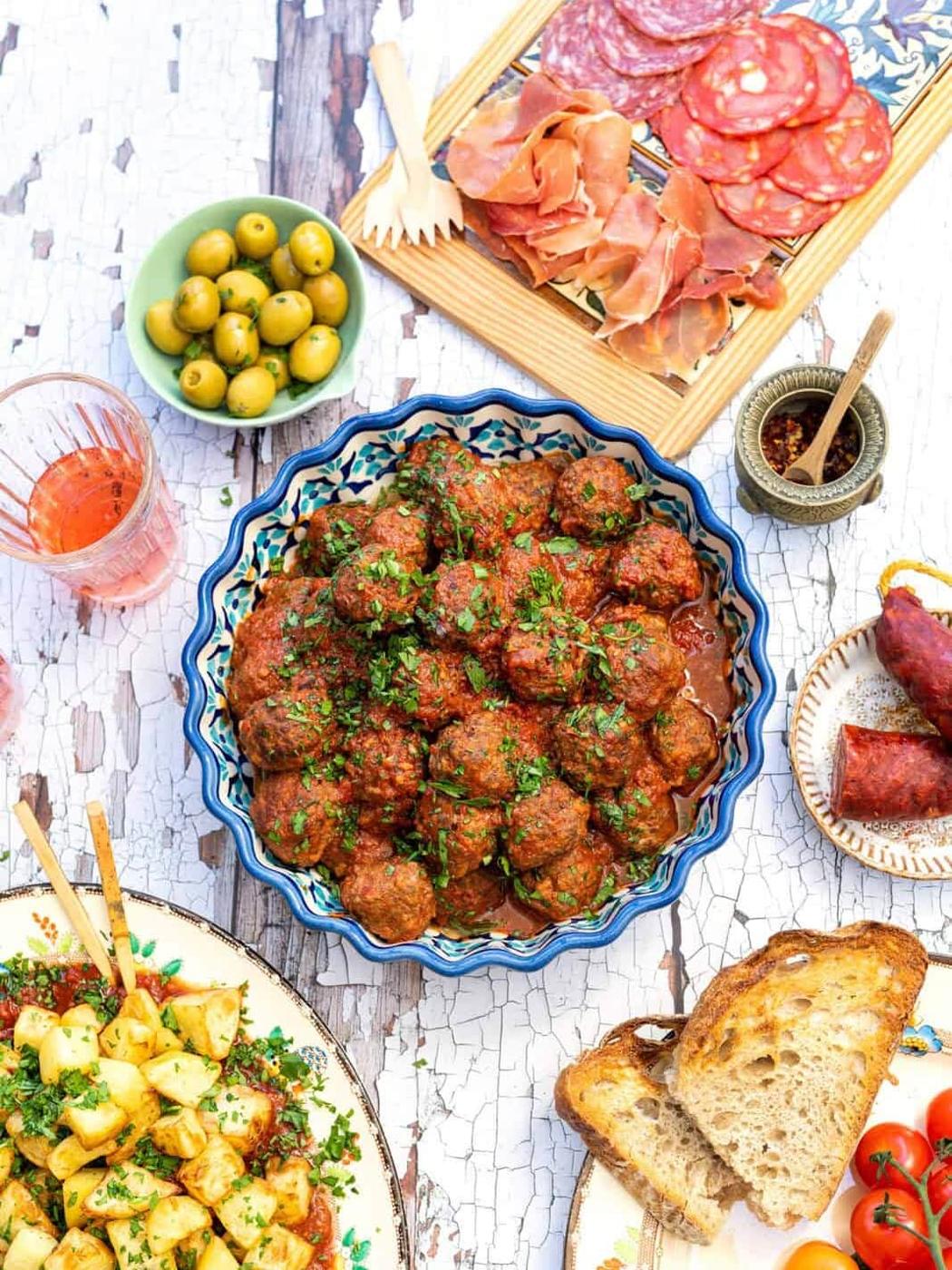 A colorful spread of Spanish tapas featuring albondigas (Spanish meatballs) in tomato sauce, cured meats, green olives, sautéed potatoes, rustic bread slices, and a glass of rosé on a rustic white table.