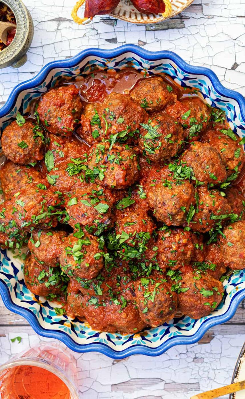 A decorative bowl filled with Spanish meatballs, or albondigas, in a rich tomato sauce, garnished with chopped fresh herbs, sits on a rustic white table.