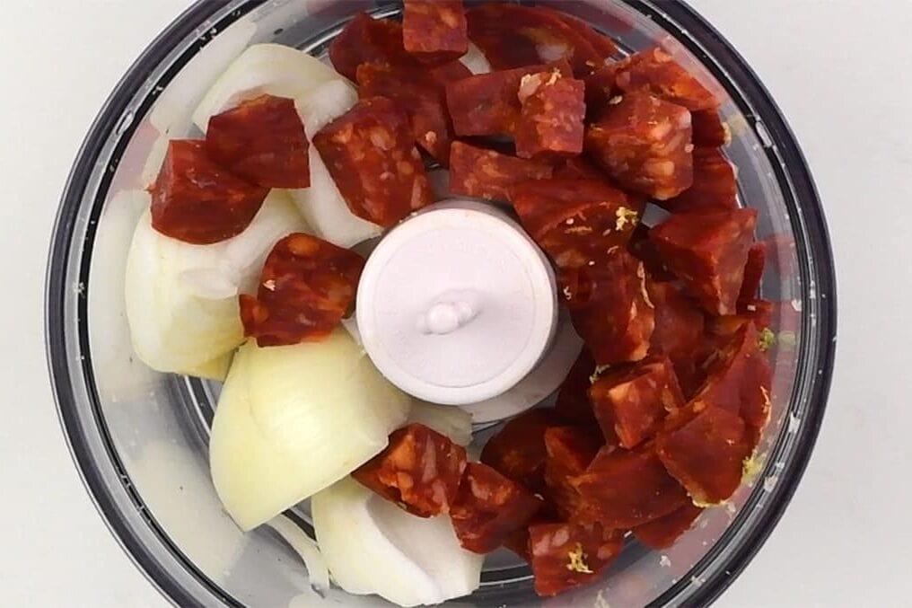 Overhead view of a food processor bowl containing chunks of onion and sliced chorizo sausage, ready to be blended for flavorful Spanish meatballs (albondigas).