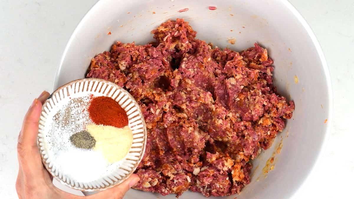 A large bowl of raw ground meat mixture sits on a white surface. A hand holds a small bowl containing measured portions of salt, pepper, paprika, garlic powder, and onion powder above the meat for making Spanish meatballs (albondigas).