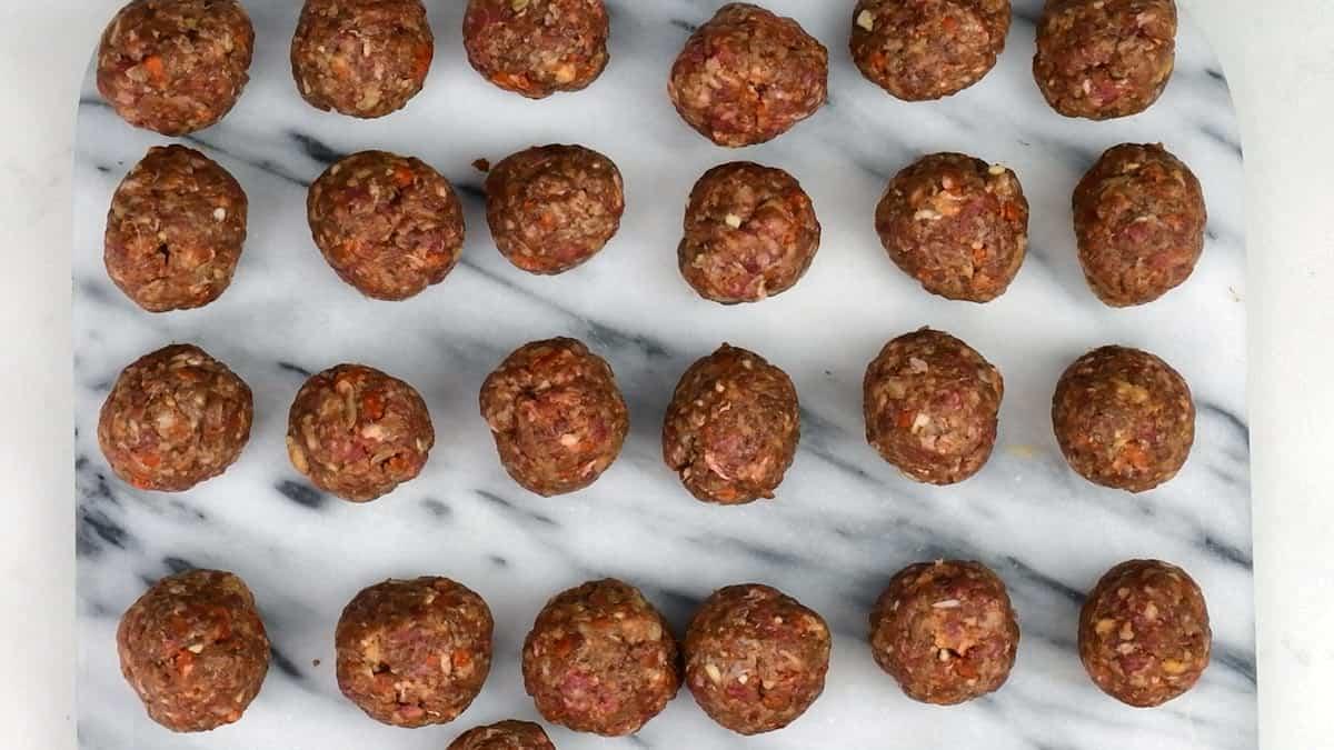Twenty-four raw albondigas, or Spanish meatballs, are arranged in neat rows on a white marble surface with gray veining. The meatballs are evenly sized and spaced apart.