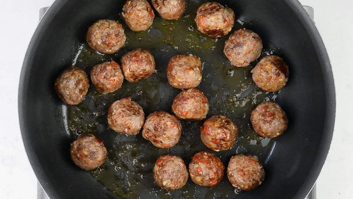 Albondigas, or Spanish meatballs, frying in oil in a black skillet, shown from above. The meatballs are browned, with some oil pooling around them in the pan.
