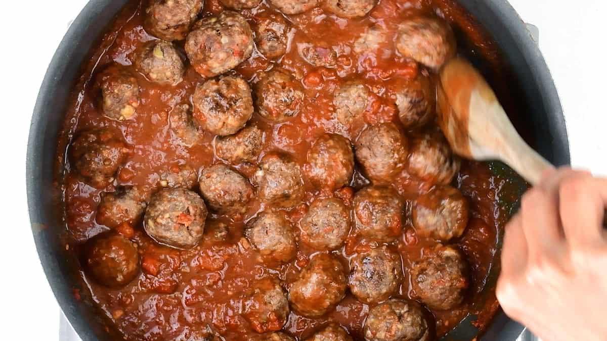 A hand stirs a pan filled with Spanish meatballs, or albondigas, simmering in a thick, chunky tomato sauce using a wooden spoon.