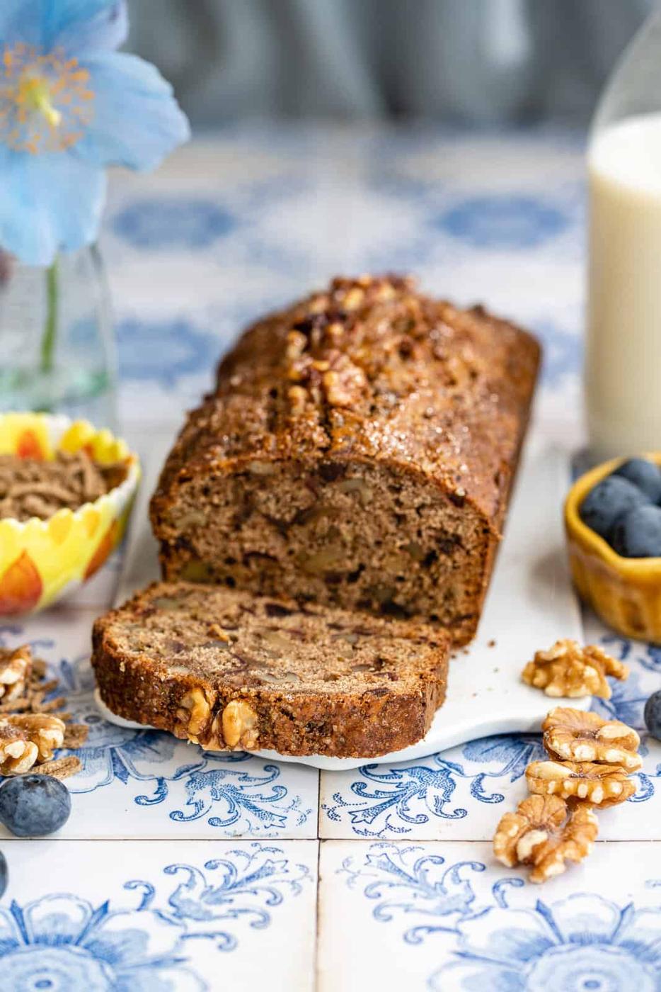 A sliced All-Bran Loaf with walnuts rests on a decorative blue and white tile, surrounded by blueberries, walnuts, a glass of milk, and a vase with a blue flower.