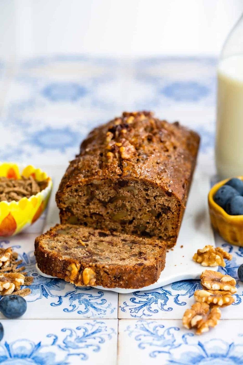A sliced All-Bran Loaf with walnuts rests on a decorative blue and white tile, surrounded by blueberries, walnuts, a glass of milk, and a vase with a blue flower.
