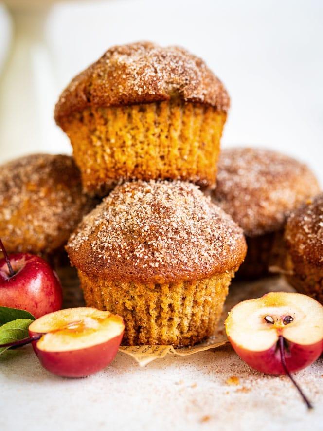 A close-up of cinnamon sugar muffins stacked with small red apples and apple halves in front, all set on a white surface. The muffins have a golden-brown, sugar-dusted top.