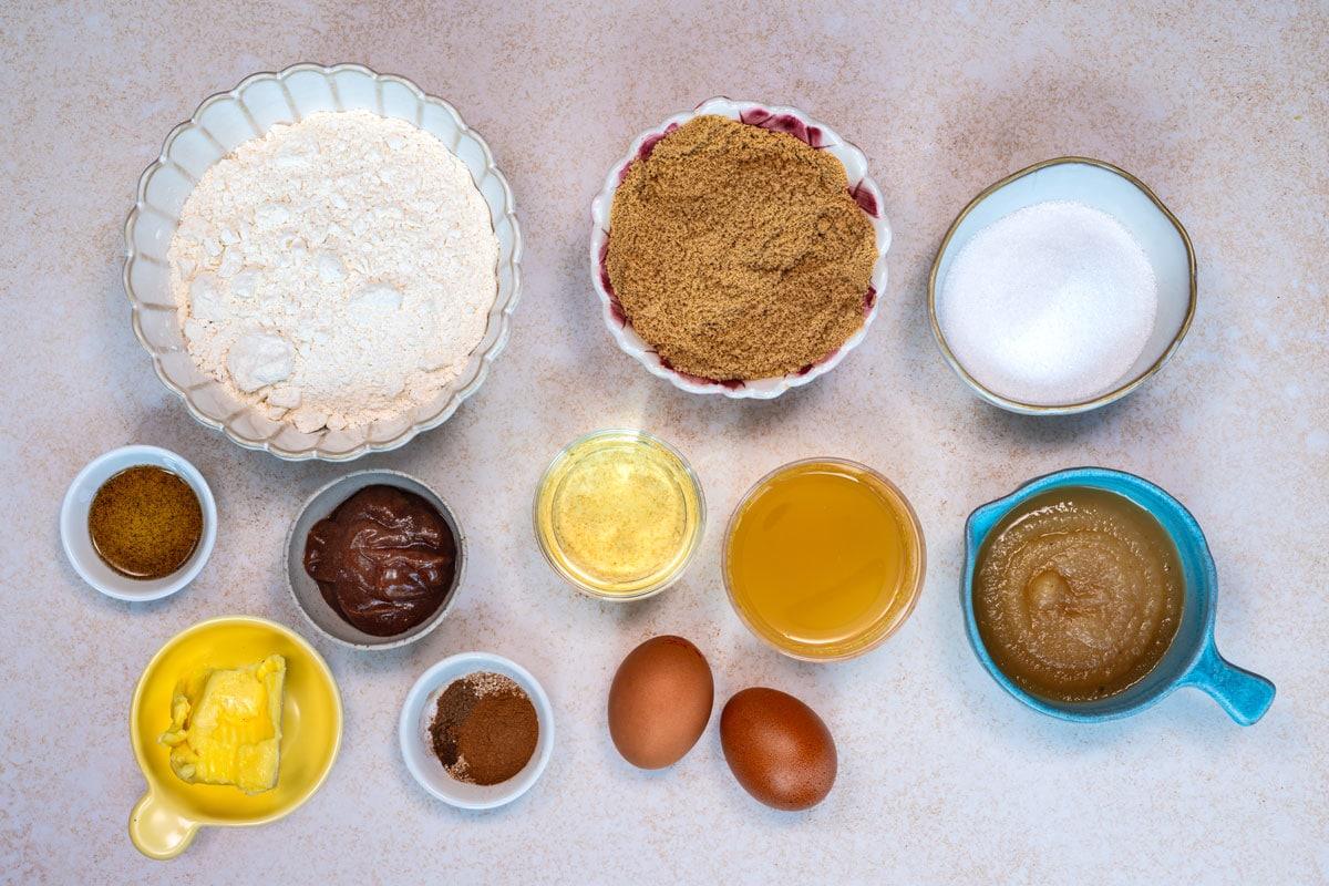A variety of baking ingredients in bowls on a countertop, including flour, brown sugar, white sugar, eggs, butter, applesauce, oil, spices, baking powder, and cocoa powder, all arranged neatly in rows.