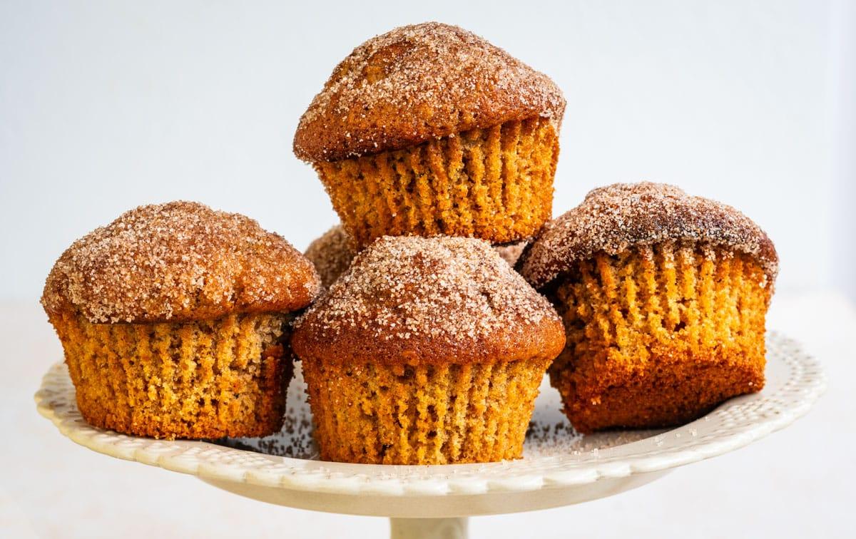 Five apple cider muffins coated with cinnamon sugar are arranged on a decorative white cake stand, with a golden-brown color and sugar topping, against a light background.