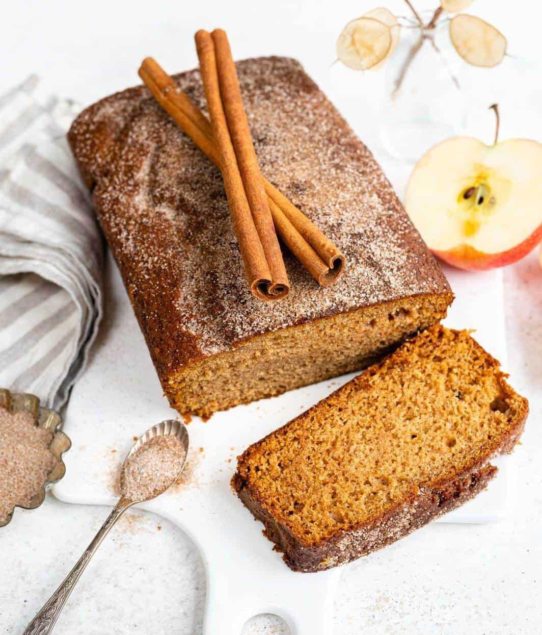 A loaf of Apple Cinnamon Bread with a sliced piece in front, topped with cinnamon sticks, sits on a white cutting board. Nearby are a spoon with sugar, a half apple, and a striped cloth napkin.