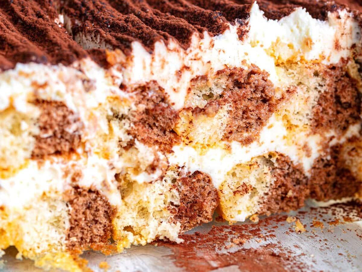 A close-up of a sliced tiramisu dessert in a glass dish, showing layers of coffee-soaked ladyfingers, creamy mascarpone filling, and cocoa powder dusted on top.