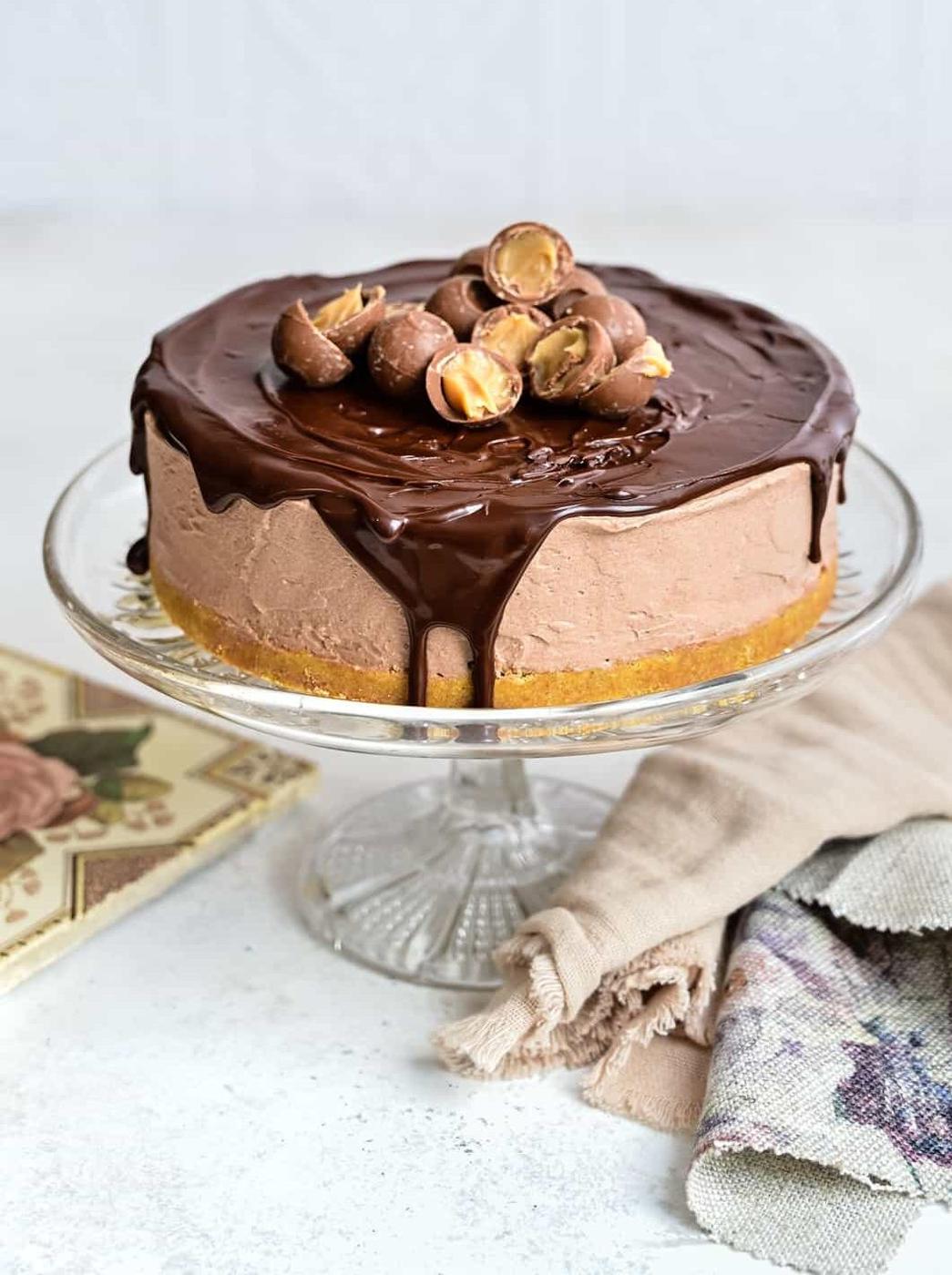 A no bake Baileys Cheesecake sits on a glass cake stand, topped with glossy chocolate ganache and halved chocolate hazelnut truffles. A folded napkin and a book rest beside the stand on a light surface.