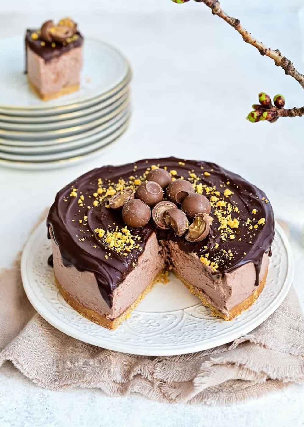 A no bake Baileys Cheesecake topped with chocolate balls and crushed nuts, with a slice removed, sits on a white plate. A stack of plates holds the removed slice in the background. A branch with buds is in the top right corner.