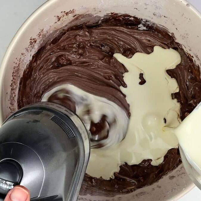 A hand holds an electric mixer blending chocolate batter in a bowl while another pours in creamy liquid, likely Baileys, perfect for a no bake Baileys cheesecake recipe.