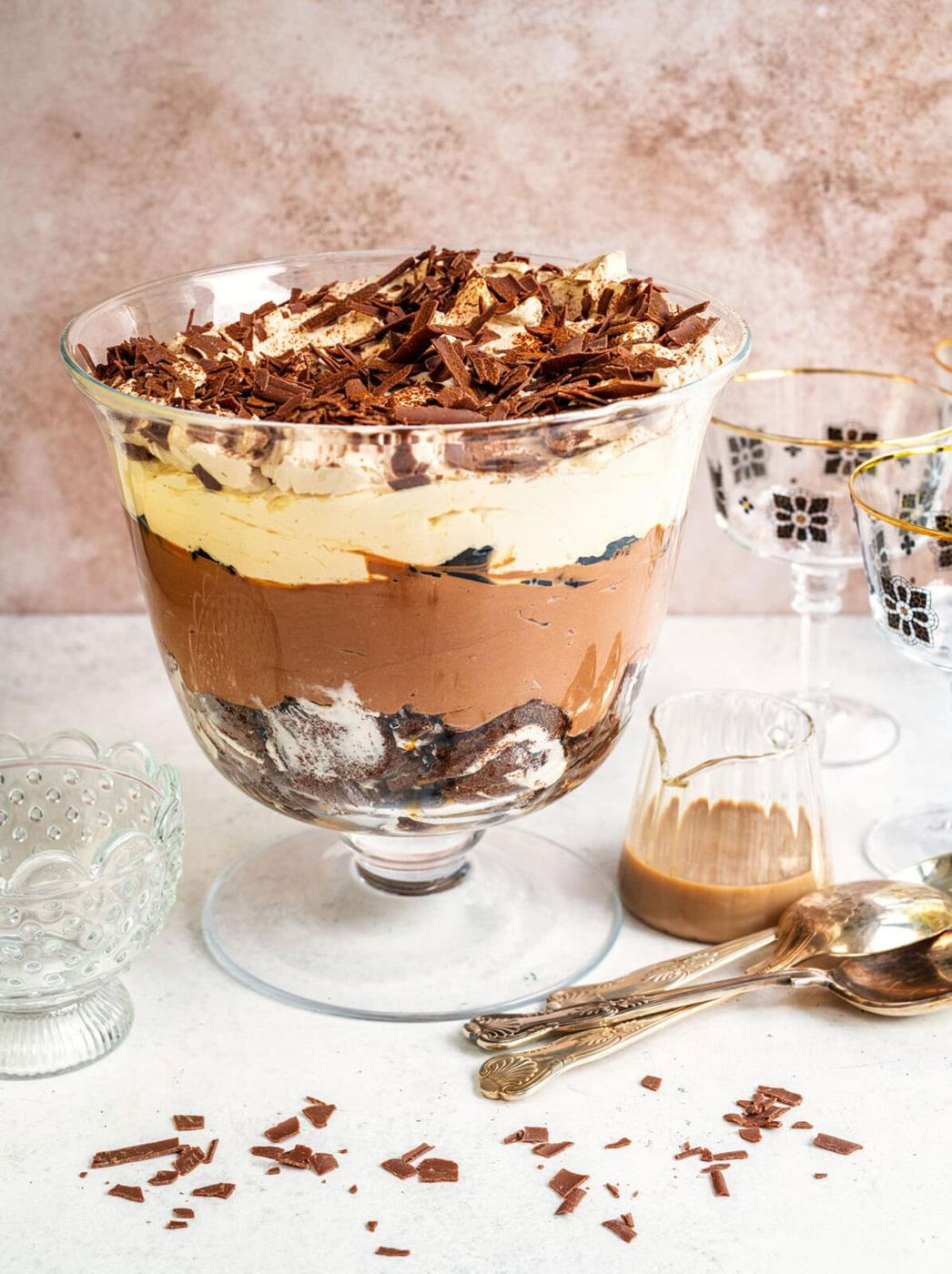 A layered chocolate trifle in a glass bowl, topped with chocolate shavings, sits on a white surface. Nearby are vintage glasses, a small jug of sauce, an empty bowl, and a few spoons with chocolate pieces scattered around.
