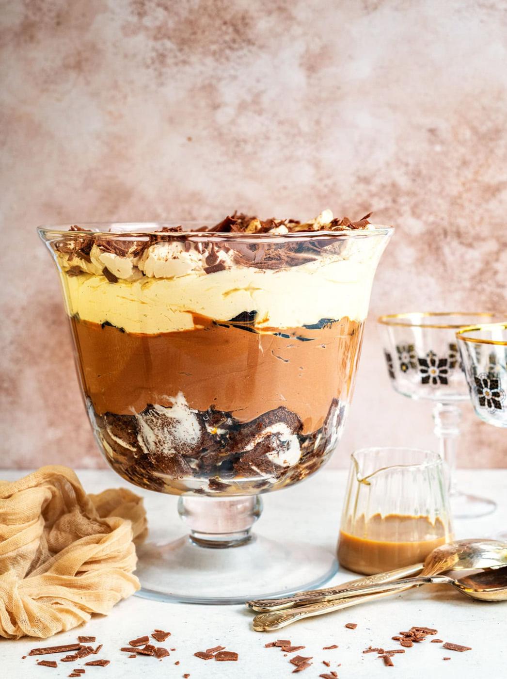 A layered chocolate and cream trifle topped with chocolate shavings, served in a glass trifle dish. Nearby are two decorative glasses, a small glass pitcher with caramel, a golden spoon, and a beige cloth.