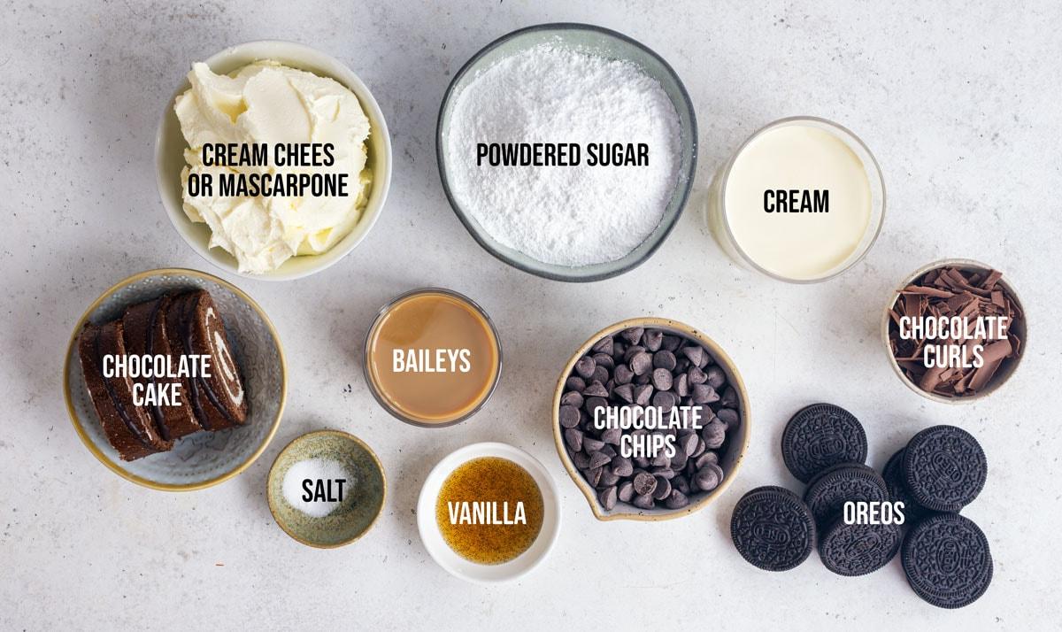 Top-down view of nine labeled bowls and cups containing ingredients for dessert: cream cheese or mascarpone, powdered sugar, cream, chocolate cake, Baileys, chocolate chips, salt, vanilla, chocolate curls, and Oreos.