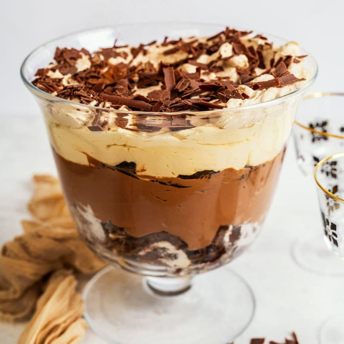 A glass trifle bowl filled with layered dessert, including whipped cream, chocolate cheesecake layer, and chocolate shavings on top. Brown and white swirled layers are visible, with chocolate pieces scattered around the base.