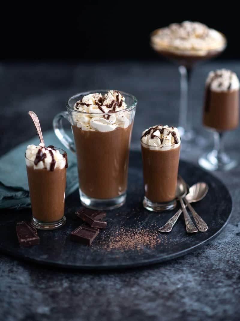 Four glasses of creamy Baileys chocolate mousse topped with whipped cream and chocolate drizzle are arranged on a dark round tray, accompanied by silver spoons and chocolate pieces. The background is dark and blurred.