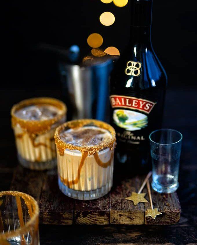A bottle of Baileys Irish Cream sits next to two creamy Baileys White Russian cocktails in glasses with caramel-drizzled rims on a rustic wooden board, with golden bokeh lights in the background. A metal shaker and empty glass are nearby.