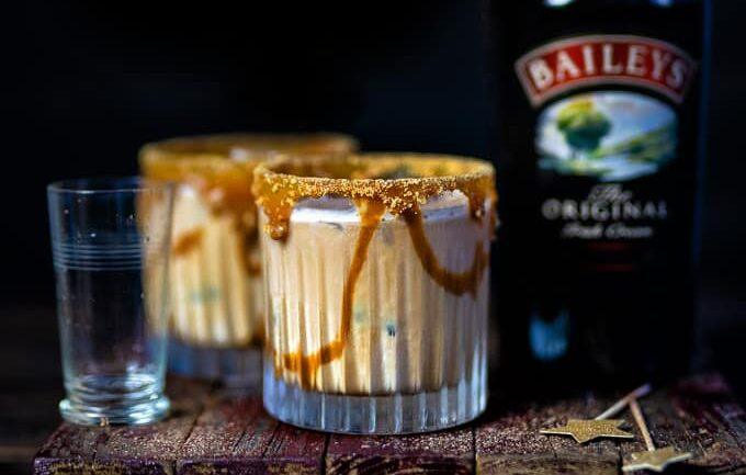 Two creamy Baileys White Russian cocktails in short glasses with caramel drizzle and sugared rims sit on a rustic wooden board next to a tall bottle of Baileys Irish Cream and a small glass of water, all against a dark background.