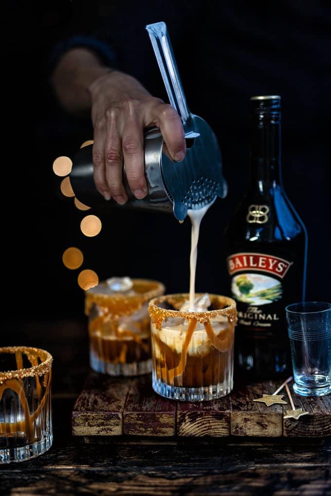 A hand pours a creamy Baileys White Russian cocktail from a shaker into a caramel-drizzled glass, next to a Baileys Irish Cream bottle and two matching drinks on a rustic wooden surface.