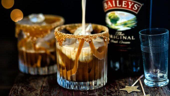 Two glasses with caramel-drizzled rims are being filled with a creamy Baileys White Russian cocktail over ice, next to a bottle of Baileys Irish Cream, an empty glass, and festive star-topped stirrers on a rustic wooden surface.