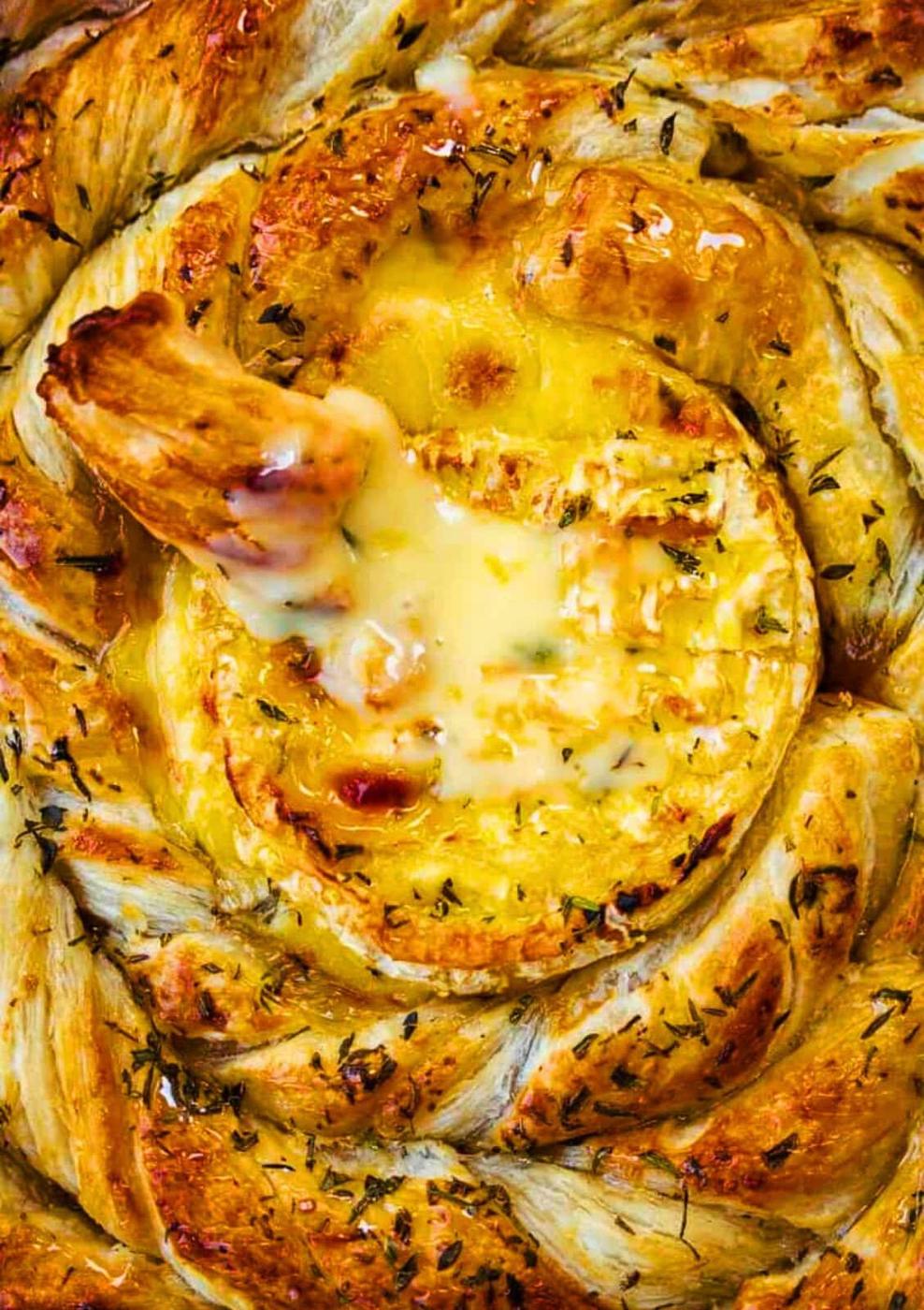 A golden, flaky puff pastry is twisted into a ring for this baked camembert recipe, filled with warm, melted cheese in the center. A piece of bread is dipped into the gooey camembert, with dried herbs sprinkled on top.