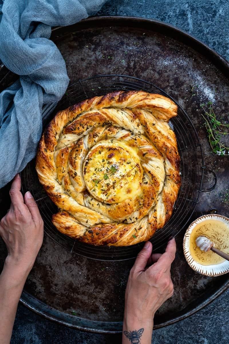 A person’s hands hold a round wire rack with a golden, braided puff pastry wreath encircling a baked camembert, sprinkled with herbs, on a rustic tray with a blue-gray cloth and honey dipper nearby—a cozy baked camembert recipe scene.