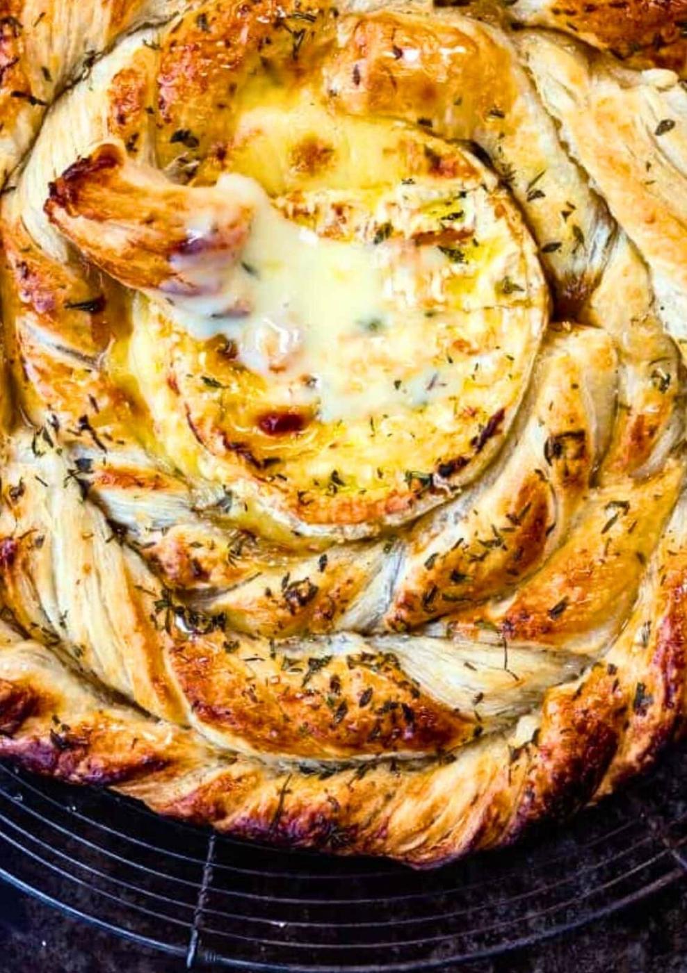 Golden, braided puff pastry surrounds a round of baked camembert, with herbs sprinkled on top and a pastry piece dipped into the gooey center—an irresistible baked camembert recipe.