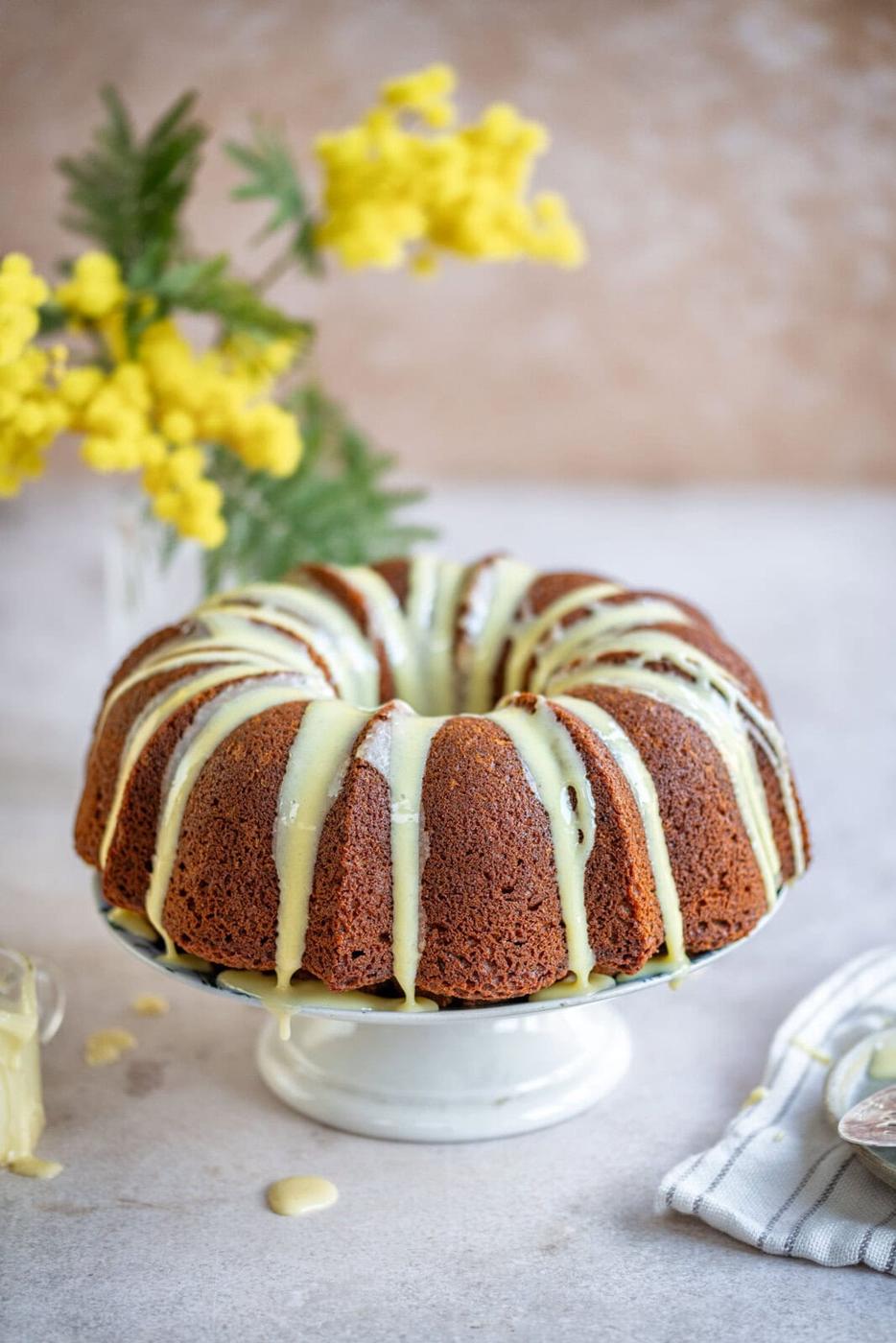A banana pudding bundt cake sits on a white stand, drizzled with pale yellow icing. In the background, a vase holds bright yellow flowers and green leaves. A striped cloth and icing bowl are nearby, all set on a light-colored surface for an inviting display.