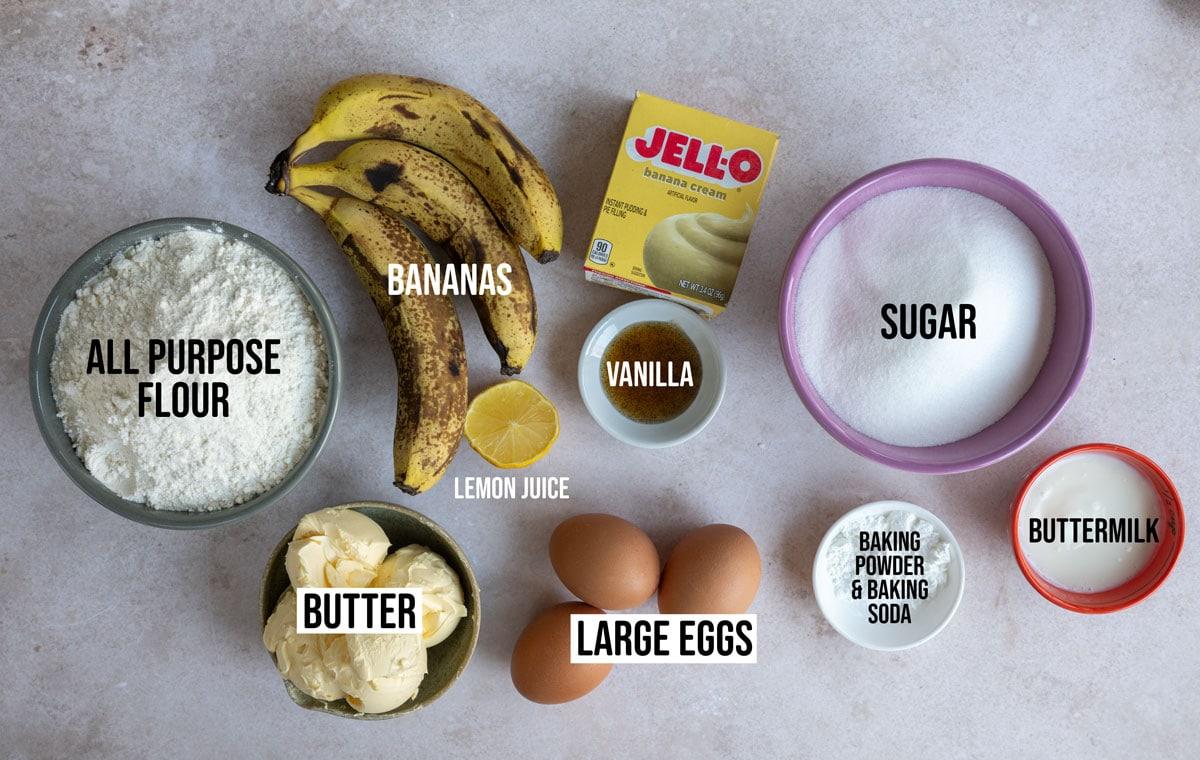 Ingredients arranged on a surface for baking: flour, butter, large eggs, sugar, bananas, banana cream pudding mix, vanilla, lemon juice, buttermilk, baking powder, and baking soda. Labels are placed on each item.