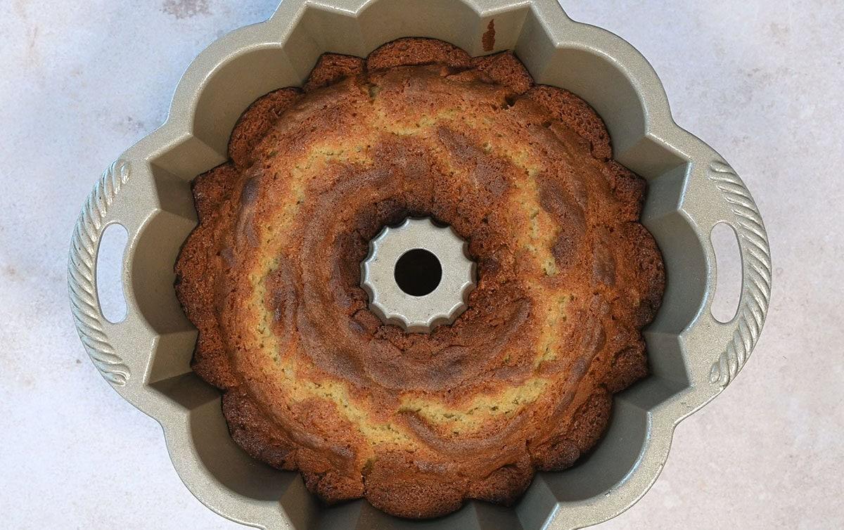 A Bundt cake with a marbled pattern, featuring lighter and darker brown swirls, is freshly baked and still in its metallic Bundt pan. The pan has decorative fluted edges and is placed on a light-colored surface.